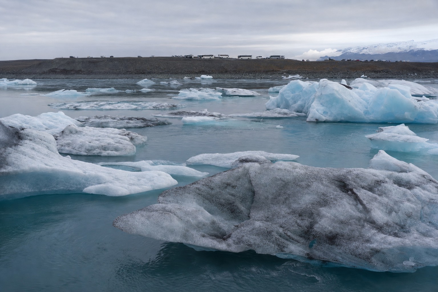Floating icebergs on Jökulsárlón glacier lagoon under soft light, with a car park and coaches small in the distance