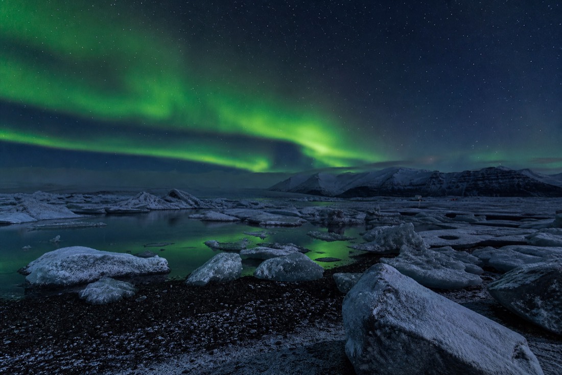 Green aurora over Jökulsárlón in winter with snow-dusted icebergs on black sand under a dark sky