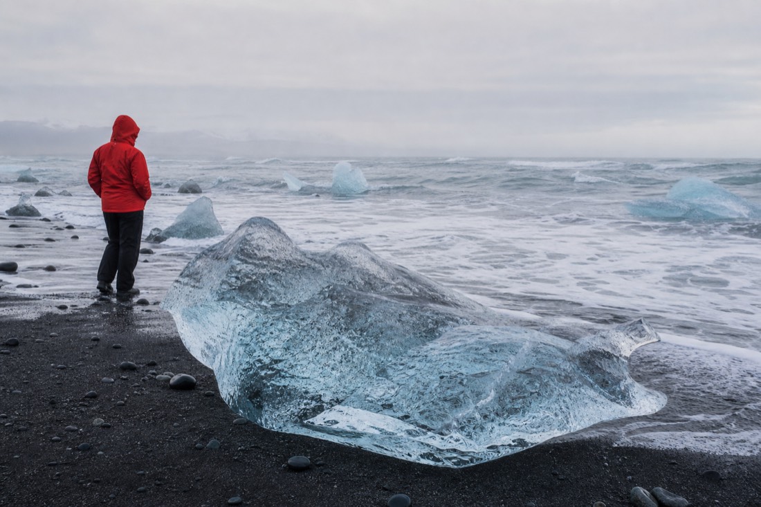 A traveller in a windproof shell jacket standing on wet black sand beside a stranded chunk of clear glacier ice