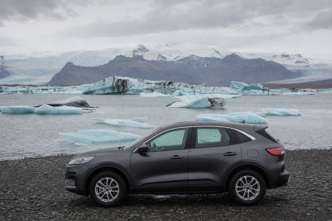 A rental car parked on the shore of Jökulsárlón Glacier Lagoon with icebergs behind it on an overcast Icelandic day