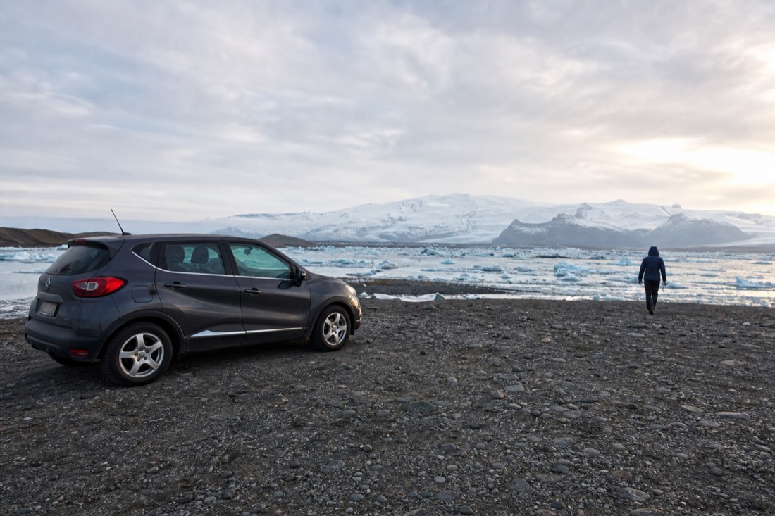 A car parked near Jökulsárlón in soft evening light while a traveller walks alone toward the lagoon shore