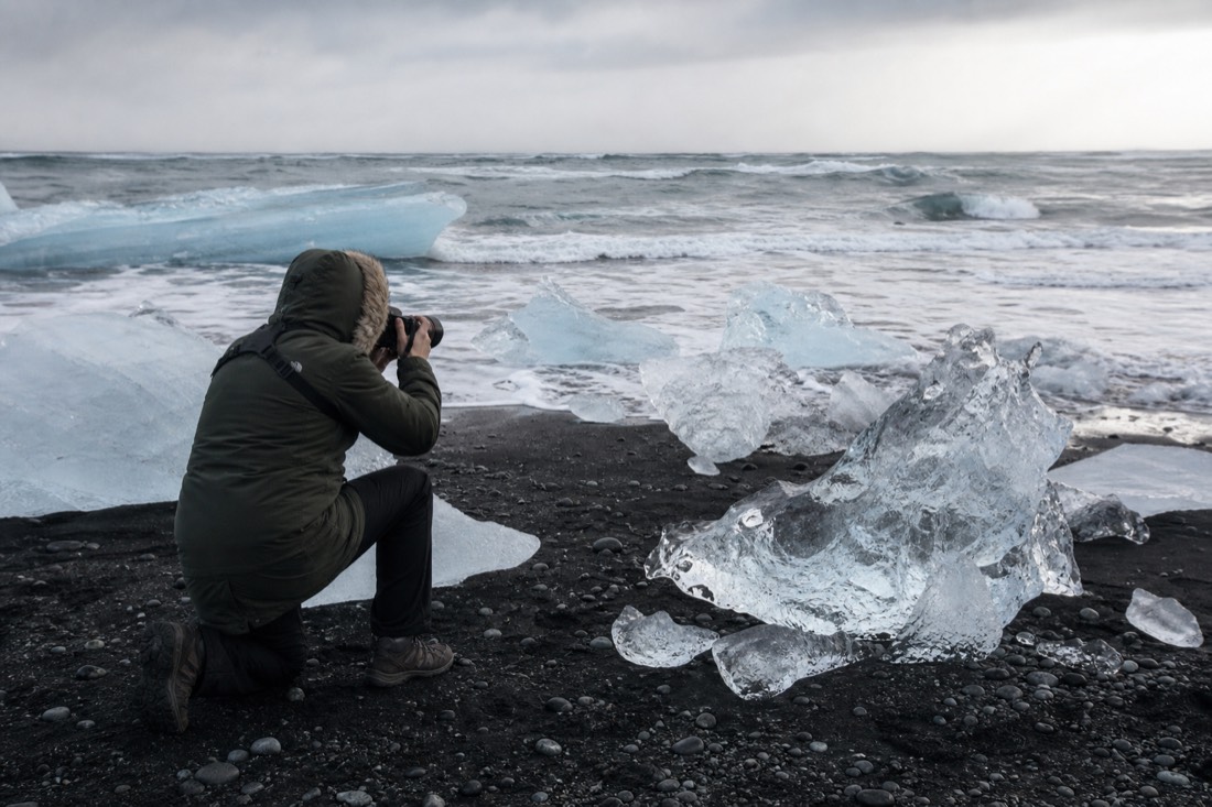 A visitor in warm waterproofs walking along the black sand of Diamond Beach photographing glacier ice, south-east Iceland