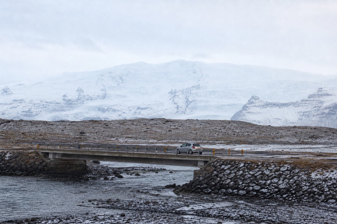 A small rental car crossing a snow-dusted single-lane bridge on Route 1 in low winter light with Vatnajökull behind on the way to Jökulsárlón